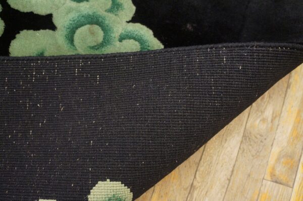 Black woven rug backing is curled to show the green floral pattern and black pile on wood flooring.