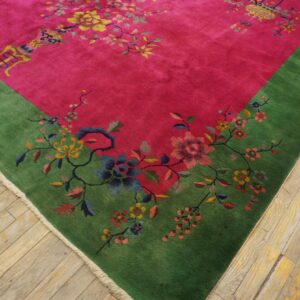 Pink rug with a wide green border and colorful floral pattern lies on distressed wooden floorboards.