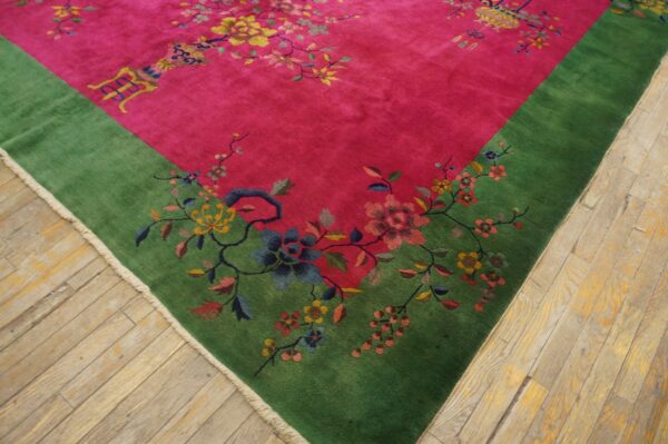 Pink rug with a wide green border and colorful floral pattern lies on distressed wooden floorboards.