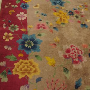 Multicolored floral pattern spans the red border and tan field of a textured rug on worn wood planks.