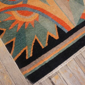 Teal, orange, and black geometric rug with striped border lying on distressed light wood planks.