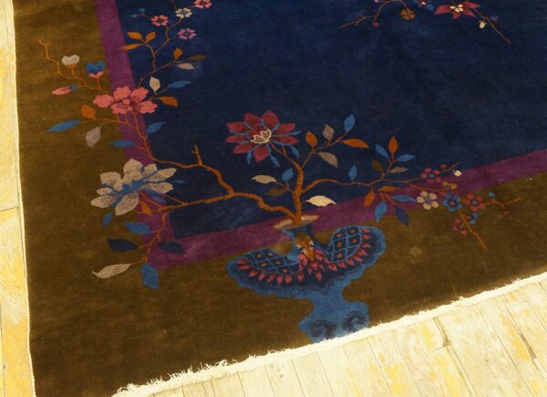 Dark blue rug with a brown border and pink floral designs centered around a stylized blue vase.