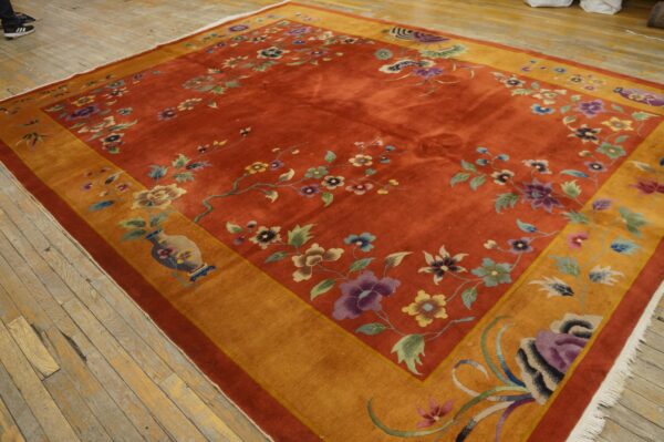 Rust-colored rug with multicolored floral design, a golden-orange border, and white fringe on wood planks.
