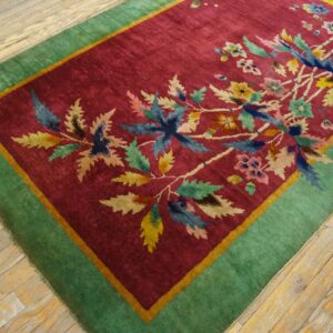 Deep red rug featuring colorful floral motifs and a wide green border on distressed light wood flooring.