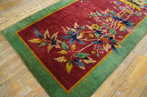 Deep red rug featuring colorful floral motifs and a wide green border on distressed light wood flooring.
