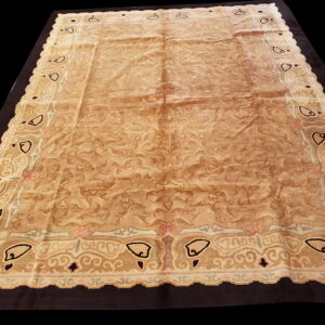 Ornate beige and brown low-pile rug with abstract border motifs displayed on a black background.