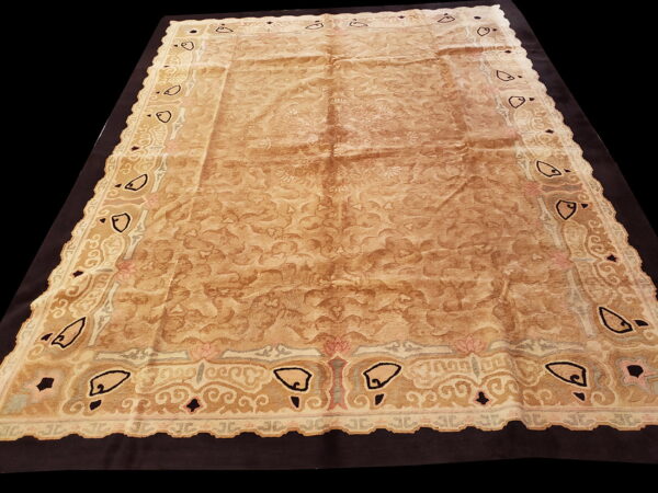 Ornate beige and brown low-pile rug with abstract border motifs displayed on a black background.