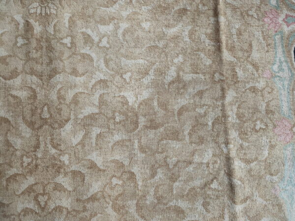 Close-up of a low-pile tan and beige rug featuring a distressed floral pattern with hints of pink and blue near the edge.