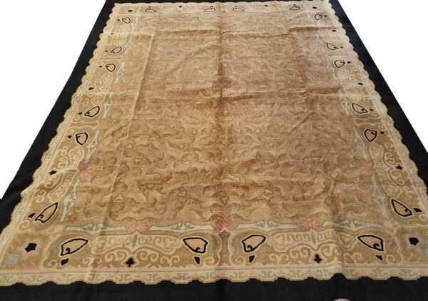 Tan floral rug with a scalloped edge border in black, rose, and pale blue, displayed on a black surface.