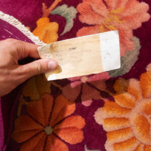 A hand holds a tag over a high-pile floral rug featuring orange flowers on a deep magenta background.