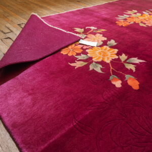 Deep magenta rug with orange floral designs partially rolled back on worn wood plank flooring.