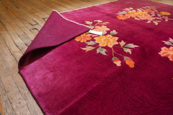 Deep magenta rug with orange floral designs partially rolled back on worn wood plank flooring.