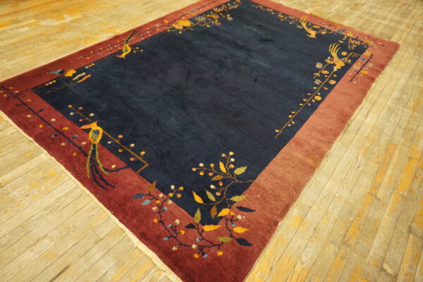 A navy blue rug with a maroon border featuring floral vine and bird motifs rests on worn wood flooring.