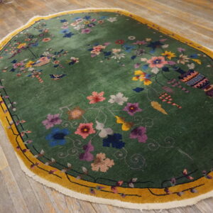 Ornate green oval rug with multicolor blossoms and a gold border sits on light rustic plank wood floors.
