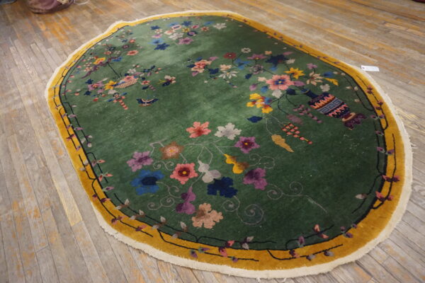Ornate green oval rug with multicolor blossoms and a gold border sits on light rustic plank wood floors.