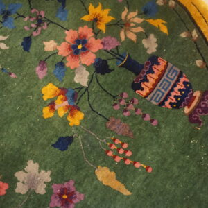 Close-up of a green rug featuring multicolored flowers, a decorative vase, and a wide yellow border.