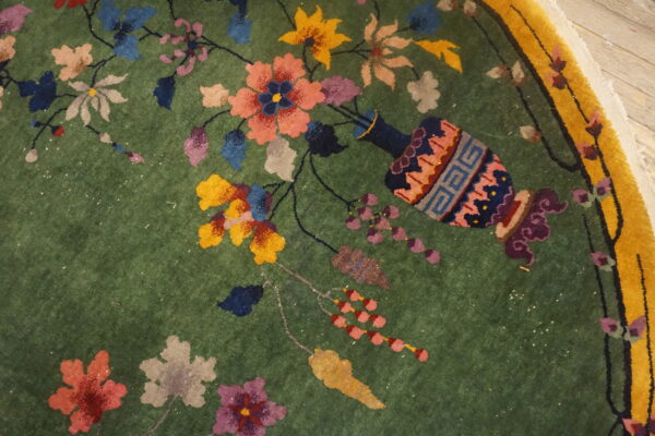 Close-up of a green rug featuring multicolored flowers, a decorative vase, and a wide yellow border.
