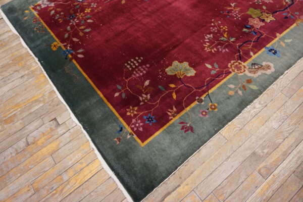 Deep red rug with multicolor floral vines and a green border lies on worn light wood flooring.