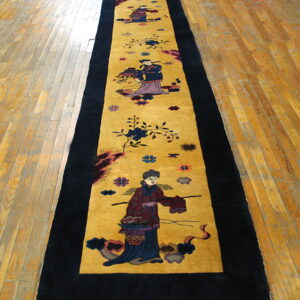 Gold runner rug with black borders and repeating figural motifs laid across worn wooden plank flooring.