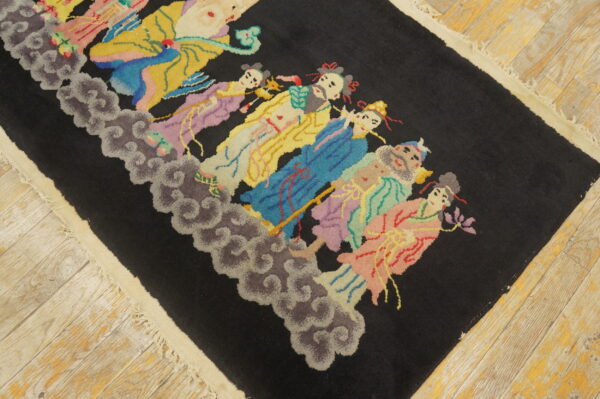 Black rug with a brightly colored figural pattern resting on distressed light wood flooring.