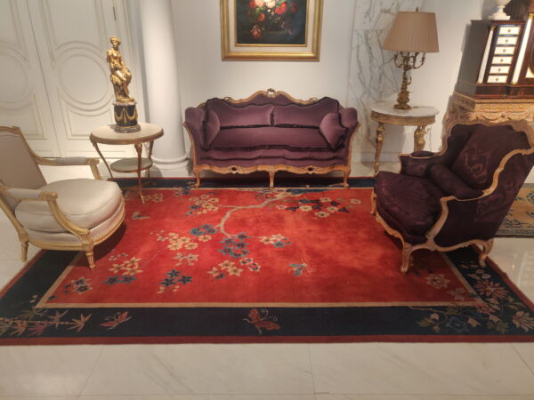 Ornate red rug with flowering branches and a navy border anchors a room with gilded furniture.