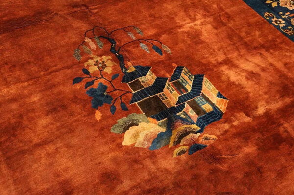 Rust rug detail showing a low pile field with a blue and brown pictorial house and floral motif.
