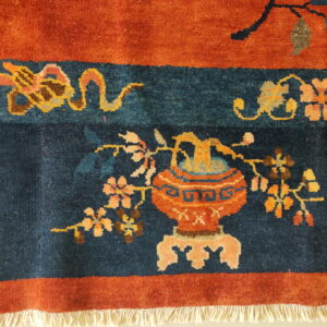 Rust and navy rug featuring an ornate vase and floral vine motifs with visible white fringe.