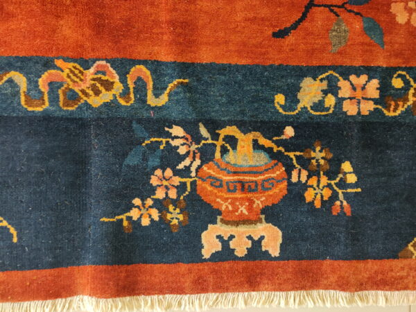 Rust and navy rug featuring an ornate vase and floral vine motifs with visible white fringe.
