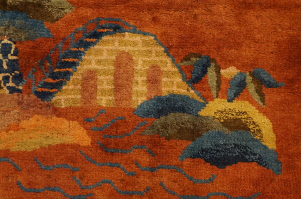 Rustic orange rug close-up with a stylized pattern of mountains and waves in blue, gold, and brown.