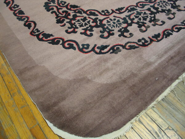 Corner of a low-pile taupe rug featuring black and red scrolling motifs rests on worn wood flooring.
