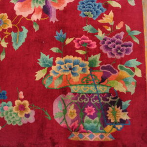 Deep red rug featuring multicolored floral and vase motifs rests beside light brown wood flooring.