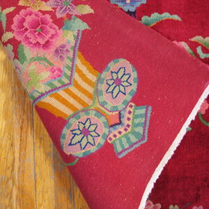 A burgundy floral rug corner folds over worn wood, revealing a pink backing with brightly woven pattern details.