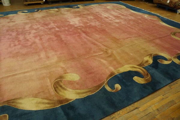 Rose-colored rug with distressed shading and a teal border featuring large scrolling gold motifs on wood flooring.