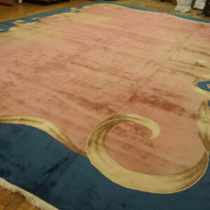 A pink low-pile rug with beige scrolling motifs along a wide teal border on wood floorboards.