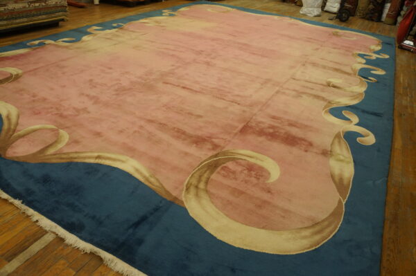 A pink low-pile rug with beige scrolling motifs along a wide teal border on wood floorboards.