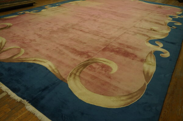 A large pink rug with a deep blue border featuring a light gold scrolling ribbon design rests on hardwood floors.