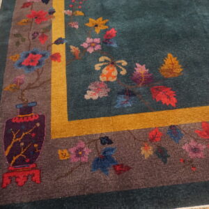 Teal rug with a colorful floral and vase pattern, brown border, and gold stripe on distressed wood floor.