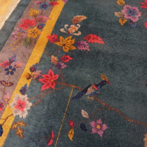 Teal rug featuring a multicolor floral and bird pattern, framed by a gold stripe, resting on wood plank flooring.