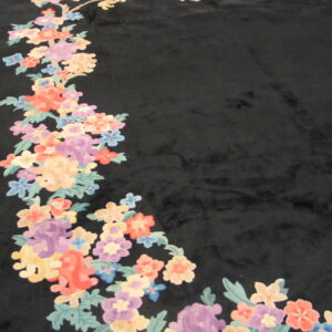 Black high-pile rug with colorful floral garland border in pastel hues rests on wood flooring.