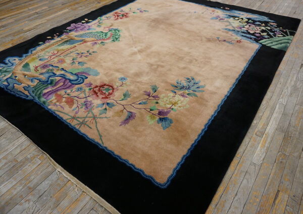 A medium-pile tan rug with vibrant floral and peacock patterns is framed by a thick black border on distressed wood.