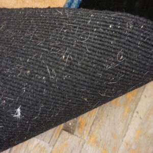 Close up of a dark, densely woven rug backing showing debris against worn wood planks.