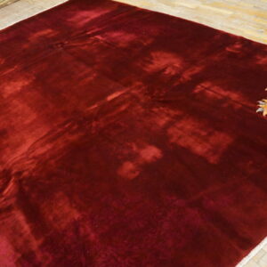 A burgundy high-sheen rug with dramatic shading and a small multicolor floral motif rests on light wood flooring.