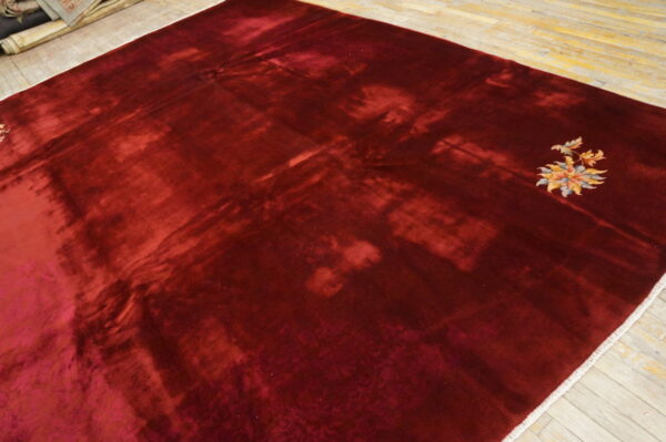 A burgundy high-sheen rug with dramatic shading and a small multicolor floral motif rests on light wood flooring.