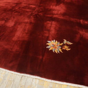 A deep maroon rug with high pile sheen and a small multi-colored floral detail on aged wood flooring.