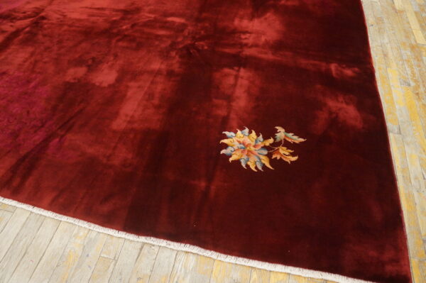 A deep maroon rug with high pile sheen and a small multi-colored floral detail on aged wood flooring.