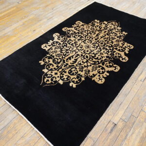A black low-pile rug with a large golden tan floral medallion rests on worn wood flooring.