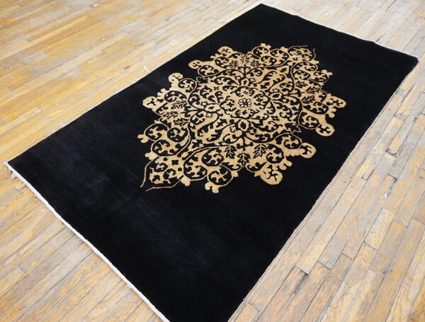 A black low-pile rug with a large golden tan floral medallion rests on worn wood flooring.