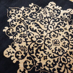 Close-up of a rug featuring a black, medium-pile background with a raised tan floral medallion pattern.