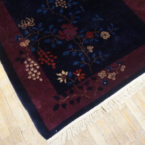 Navy floral rug with a burgundy border, white fringe, and red/tan accents rests on wood flooring.