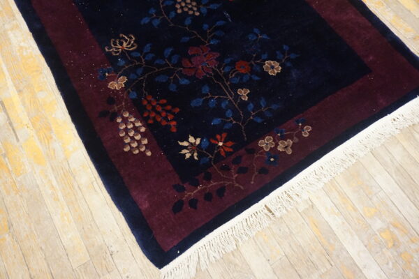 Navy floral rug with a burgundy border, white fringe, and red/tan accents rests on wood flooring.